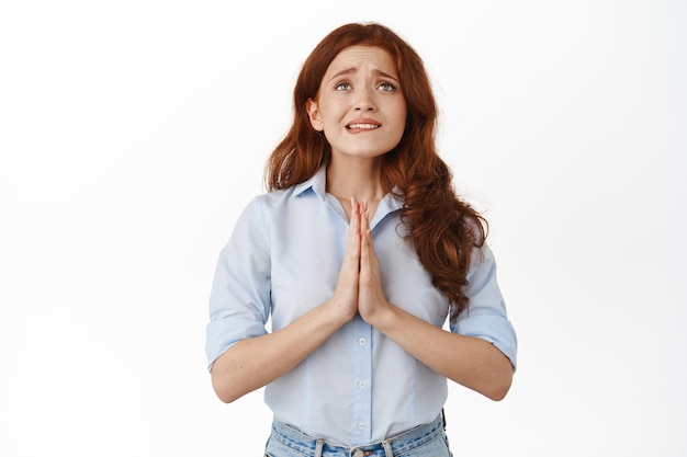 Bezorgde roodharige vrouwelijke ondernemer die god bidt, nerveus op de lip bijt en handen vasthoudt om te bidden, opkijken, smeken, wensen doen, in blouse tegen witte achtergrond staan