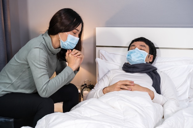 Bezorgde vrouw zorgt voor haar zieke man terwijl hij thuis op bed slaapt, mensen moeten een medisch masker dragen dat beschermt tegen pandemie van het coronavirus