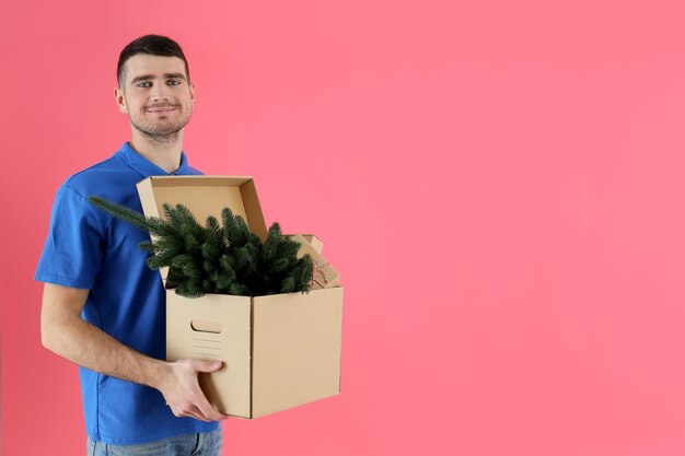 Bezorger houdt doos met kerstboom en geschenken op roze achtergrond