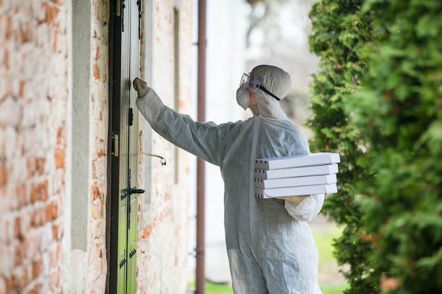 Bezorger in beschermend pak met een pizza in de buurt van een deur