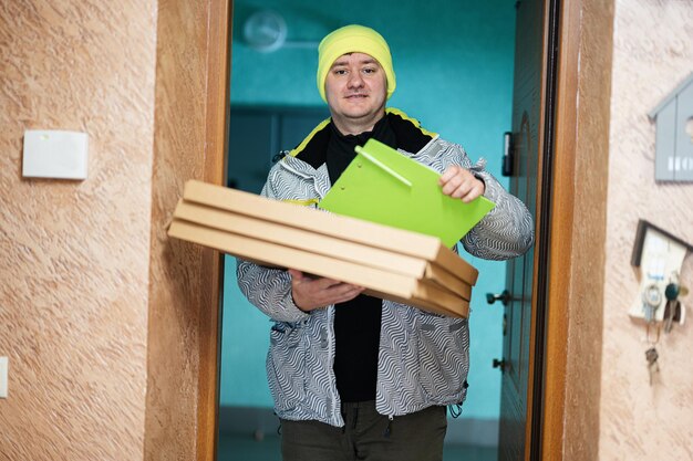 Bezorger met kartonnen pizzadozen Koerier in groene hoed die een klembord vasthoudt terwijl hij tegen de deur van een woonhuis staat