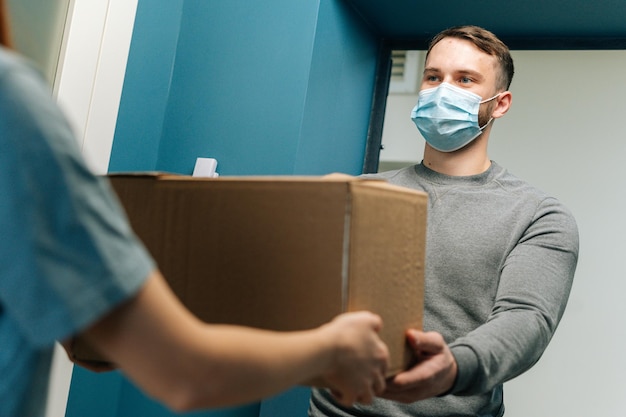 Bezorger met medisch gezichtsmasker die een kartonnen doospakket geeft aan vrouwelijke klant voor de deur thuis