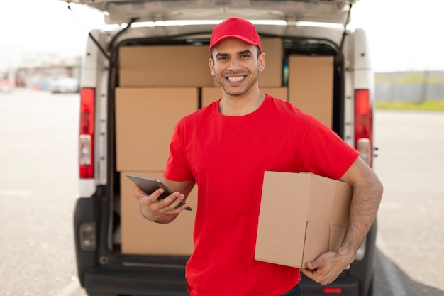 Bezorger met tablet en doos staat bij busje en glimlacht naar camera, logistiek en koerierconcept