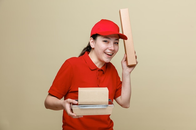 Bezorger vrouw werknemer in rode dop en blanco t-shirt uniform met voedsel dozen kijken camera gelukkig en positief glimlachend vrolijk staande over bruine achtergrond