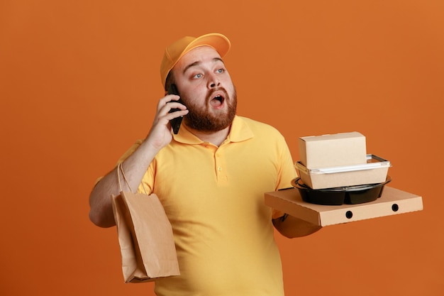 Bezorger werknemer in gele dop blanco t-shirt uniform met voedselcontainer en pizzadoos met papieren zak praten op mobiele telefoon en verbaasd kijken over oranje achtergrond