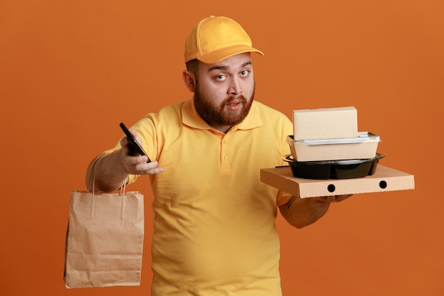 Bezorger werknemer in gele dop leeg t-shirt uniform met voedselcontainer en pizzadoos met papieren zak met smartphone die er verward uitziet over oranje achtergrond