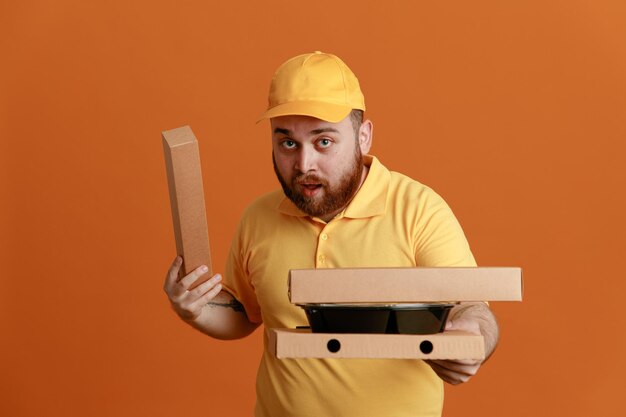 Bezorger werknemer in gele pet leeg t-shirt uniform met voedselcontainers en pizzadoos kijkend naar camera verrast over oranje achtergrond