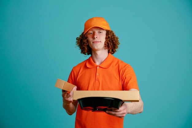 Bezorger werknemer in oranje pet leeg t-shirt uniform met pizzadoos en voedselcontainers kijkend naar camera glimlachend zelfverzekerd over blauwe achtergrond