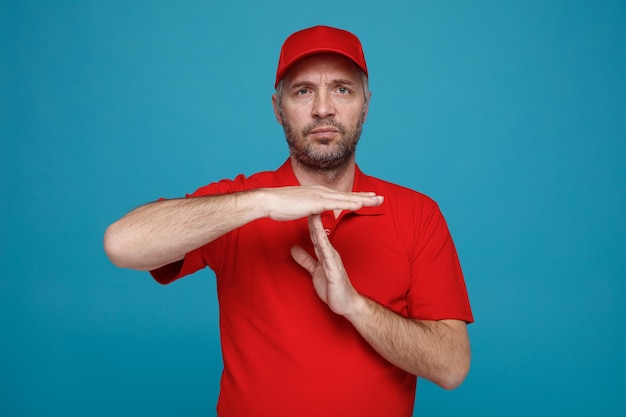 Bezorger werknemer in rode dop leeg t-shirt uniform kijken camera met fronsend gezicht time-out gebaar maken met handen permanent over blauwe achtergrond