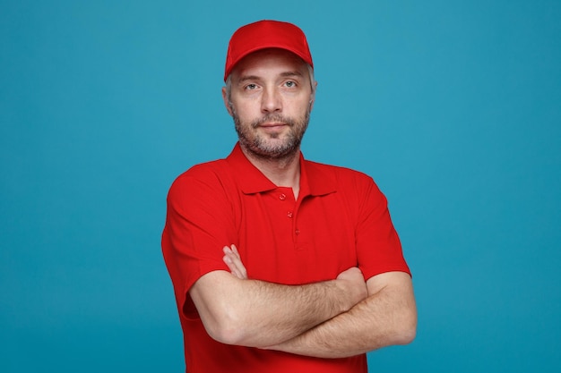 Bezorger werknemer in rode dop leeg t-shirt uniform kijken camera met gekruiste armen op zijn borst met zelfverzekerde uitdrukking staande over blauwe achtergrond