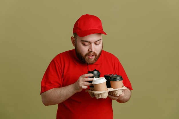 Bezorger werknemer in rode dop leeg t-shirt uniform met koffiekopjes kijken naar hen opgewonden staan over groene achtergrond