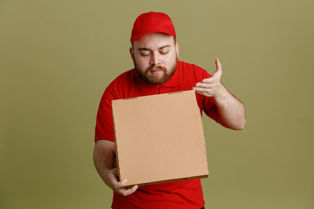 Bezorger werknemer in rode dop leeg t-shirt uniform met pizzadoos opening doos inademen van pleziergeur staande over groene achtergrond