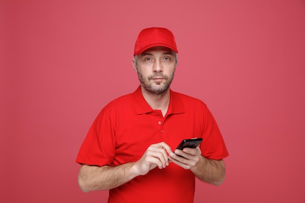 Bezorger werknemer in rode dop leeg t-shirt uniform met smartphone kijkend naar camera met serieus gezicht staande over rode achtergrond