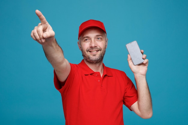 Bezorger werknemer in rode dop leeg t-shirt uniform met smartphone wijzend met wijsvinger naar de zijkant glimlachend vrolijk staande over blauwe achtergrond