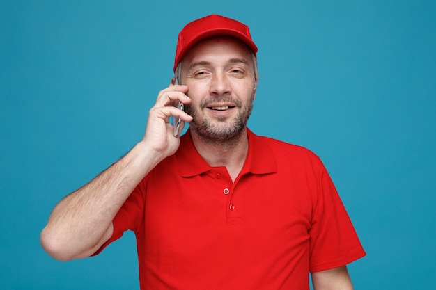 Bezorger werknemer in rode dop leeg t-shirt uniform praten op mobiele telefoon blij en positief glimlachend staande over blauwe achtergrond