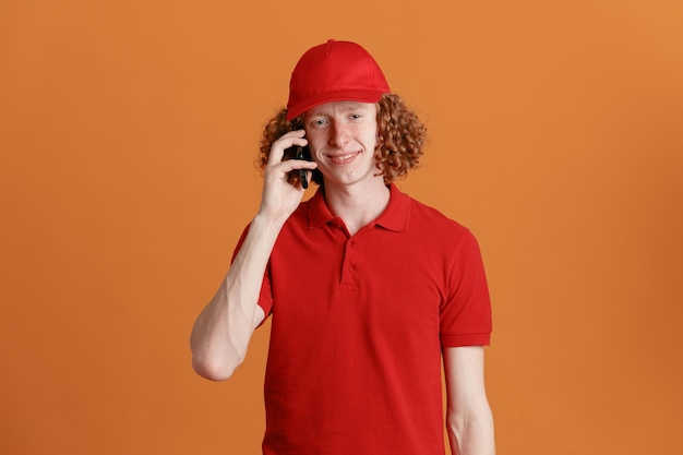 Bezorger werknemer in rode dop leeg t-shirt uniform praten op mobiele telefoon glimlachend zelfverzekerd gelukkig en positief staande over oranje achtergrond