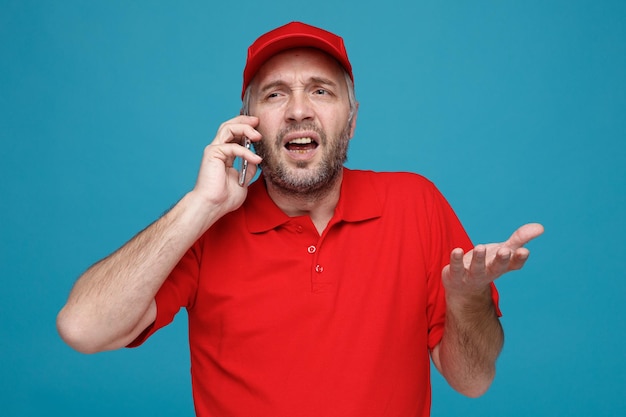 Bezorger werknemer in rode dop leeg t-shirt uniform praten op mobiele telefoon kijken verward opgeheven arm in ongenoegen staande over blauwe achtergrond