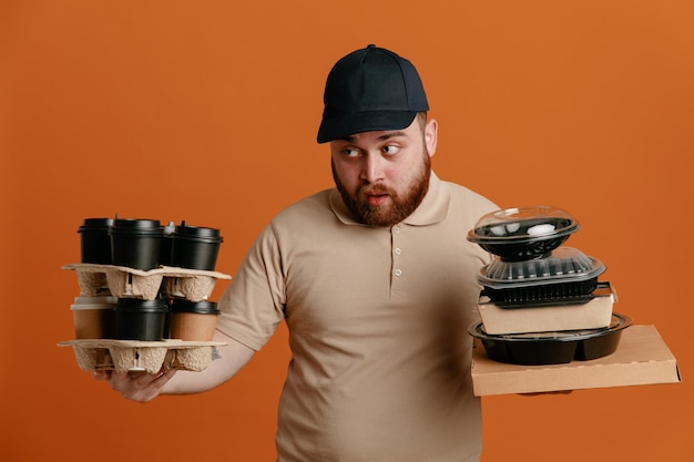 Bezorger werknemer in zwarte pet en blanco t-shirt uniform met koffiekopjes en voedselcontainers die er verward uitzien over oranje achtergrond