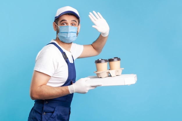 Bezorgservice. Beminnelijke koerier in overall en masker met koffie en pizzadoos, veiligheidshandschoenen dragend die drankjes eten aanbieden en hallo zwaaien naar de camera. binnen schot geïsoleerd op blauwe achtergrond