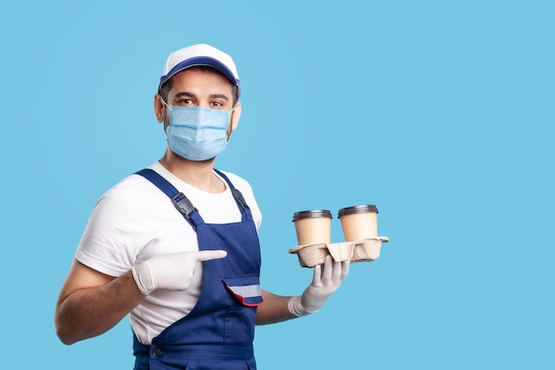 Bezorgservice. Fijne vriendelijke koeriersman in overall en masker wijzend op koffie, veiligheidshandschoenen dragend die drankjes in wegwerpbekers aanbieden en met een glimlach naar de camera kijken. binnen schot, geïsoleerd