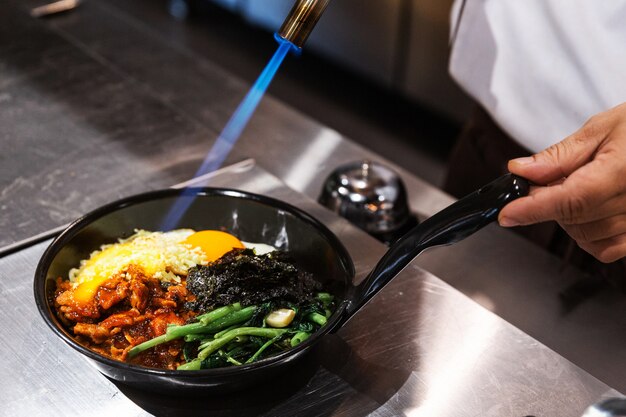 Foto bibimbap (koreaanse rijst gemengd met kimchi-varkensvlees, zeewier en roergebakken groenten met sesam) geserveerd in de hete pan die chef-kok met een zaklamp voor het smelten van mozzarella-kaas op een gebakken ei.