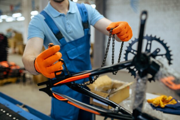 Bicycle factory, worker holds bike chain at assembly line. Male mechanic in uniform installs cycle parts in workshop