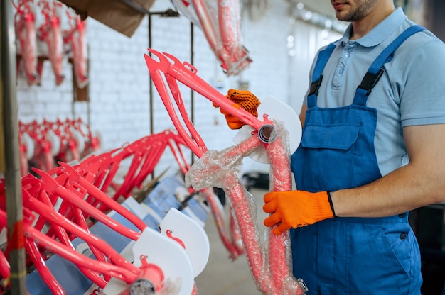 Bicycle factory, worker holds pink kid's bike frame. Male mechanic in uniform installs cycle parts, assembly line in workshop