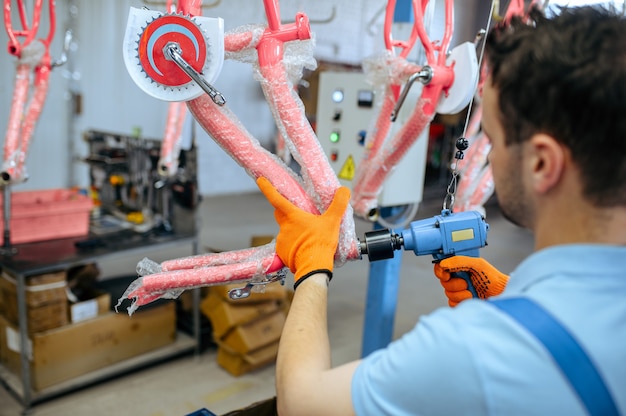 Bicycle factory, worker holds pink kid's bike. Male mechanic in uniform installs cycle parts, assembly line in workshop