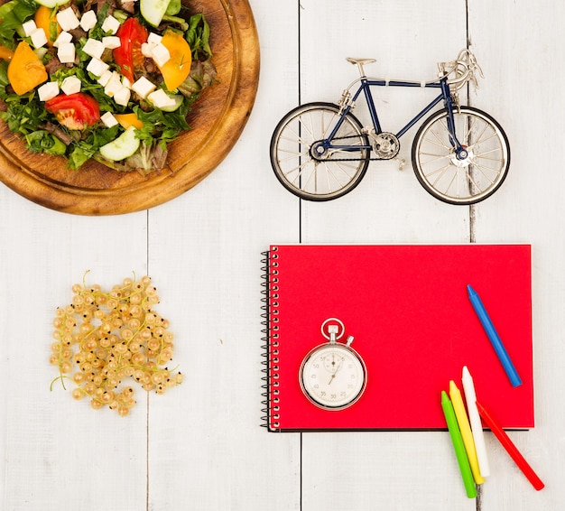 Велосипедный салат из свежих овощей, красный блокнот, секундомер и смородина на белом деревянном столе