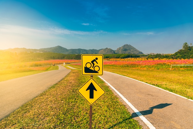 Знак велосипеда к крутой дороге с предпосылкой горной цепи и голубого неба.