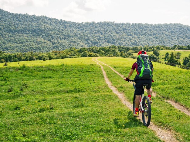 bicyclist with backpack riding mountain bike on path at green meadows background