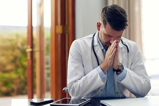 Bidden voor een succesvolle operatie Bijgesneden opname van een knappe mannelijke arts die bidt terwijl hij in zijn kantoor zit