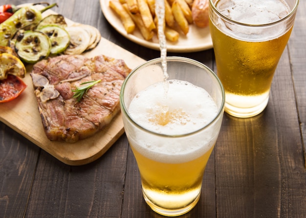 Bier wordt gegoten in glas met gastronomische steak en frietjes op houten achtergrond