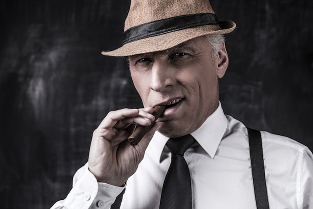 Photo big boss. serious senior man in hat and suspenders smoking cigar and looking at you while standing against dark background