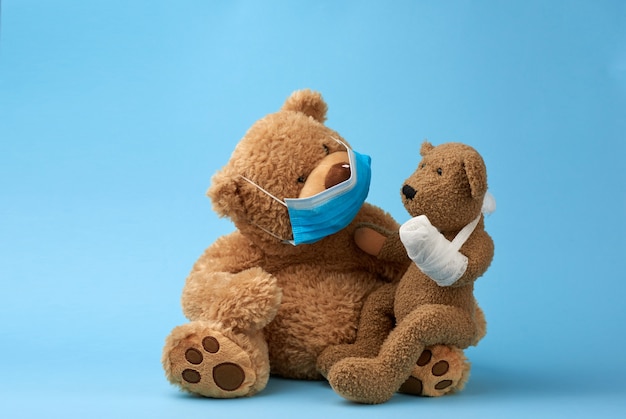 Big brown teddy bear sits in a medical mask, in his hands he holds a small toy