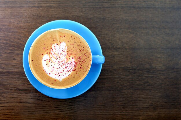 木製のテーブルにミルクと大きなコーヒーカップ。カプチーノまたはラテドリンク、茶色のテーブルにコーヒーを一杯フラットレイビュー。カフェオレのカップ。ミルクの絵やラテアート。青いマグカップのホットコーヒー