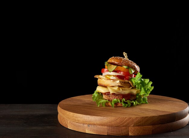 Big delicious burger with meat cutlet, cheese, fried egg, tomatoes, cucumber slices and green lettuce, fast food on a round wooden board, black background, copy space