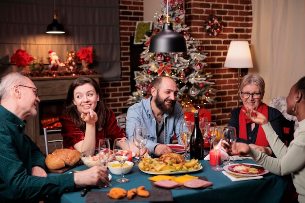 クリスマスを祝い、お祝いの夕食の席で話し、伝統的な冬の休日の食べ物を食べる多様な大家族。美しい装飾が施された場所、モミの木、背景に暖炉でクリスマスのお祝い