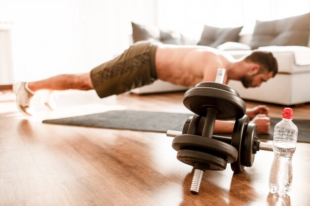 Big dumbbells and water on floor. Young man doing sport at home. Cut view of t-shirtless guy sportsman with a sports figure  in plank position.