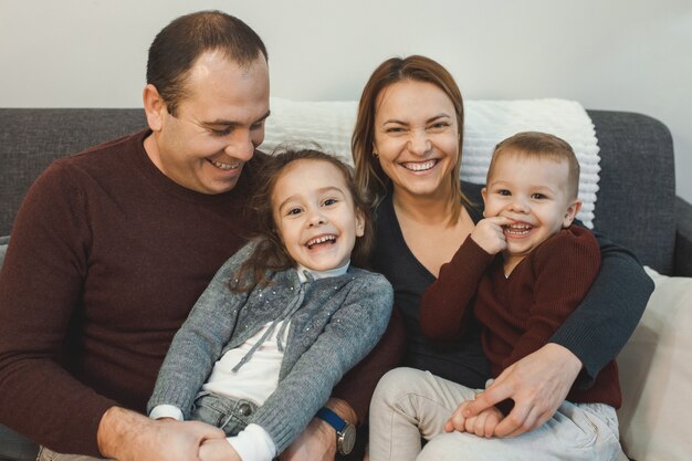 Big family portrait sitting on the couch while mother and father holding on their legs their son and daughter laughing at home.