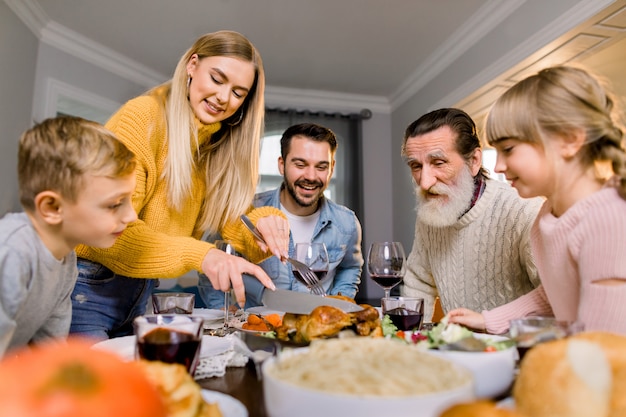 感謝祭のディナーを食べて幸せな大家族。ダイニングテーブルの七面鳥の丸焼き。お祝いの食事を持っている親子。かなりの母親が肉を切る。