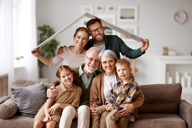 Big happy family grandparents mother father with little kids\
son and daughter celebrating relocation in new home sitting on the\
coach under paper roof and smiling at camera mortgage loan\
concept