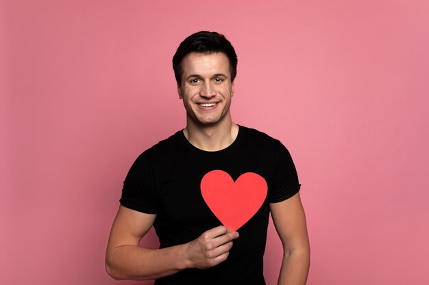 Big heart. charming man with a charming smile, who is looking in the camera and holding a red heart-shaped card.