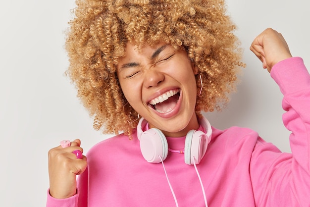 Big luck concept optimistic young woman with curly bushy hair\
clenches fists gets joy from victory wears pink jumper headphones\
around neck isolated over grey background excited over special\
offer