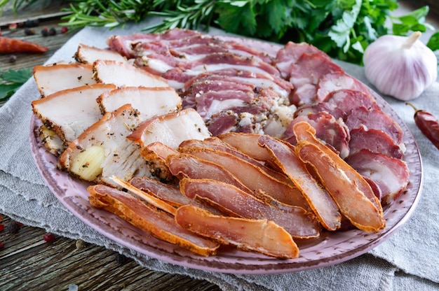 Большой мясной набор. Домашняя копченая свино-говяжья колбаса, соленый бекон, нарезанные кусочки бастурмы на деревянной доске со специями и зеленью.
