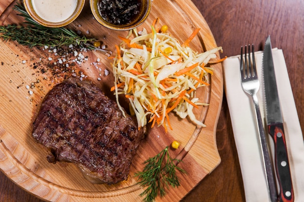 Big meat steak with vegetables and sauces, knife and fork on wooden table in luxury restau