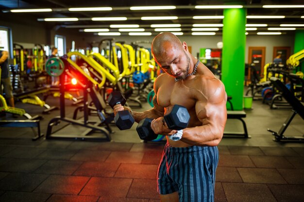 Big muscular man with a naked torso with dumbbells in his hands. Man in the gym shakes biceps