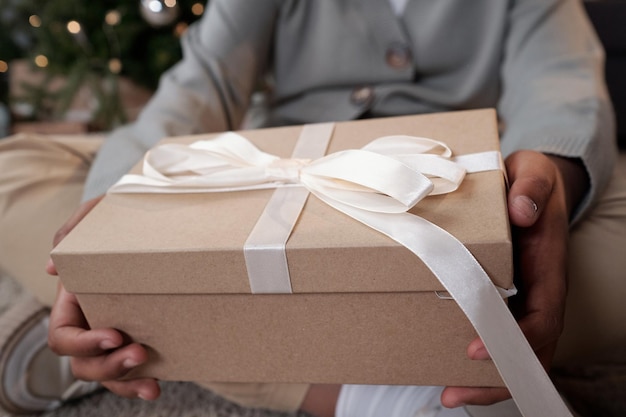 Big packed giftbox with white silk ribbon on top containing xmas present held by girl passing it to someone