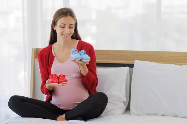 Big pregnant woman happily plays with the baby's shoes on bed in bedroom