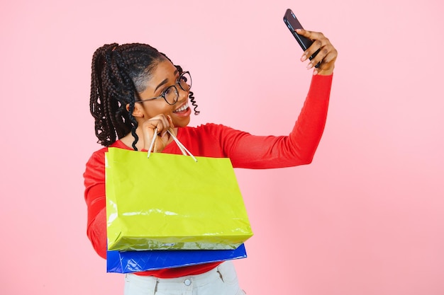 Big Sale Excited afro girl with shopping bags touching sunglasses looking at camera over pink studio wall